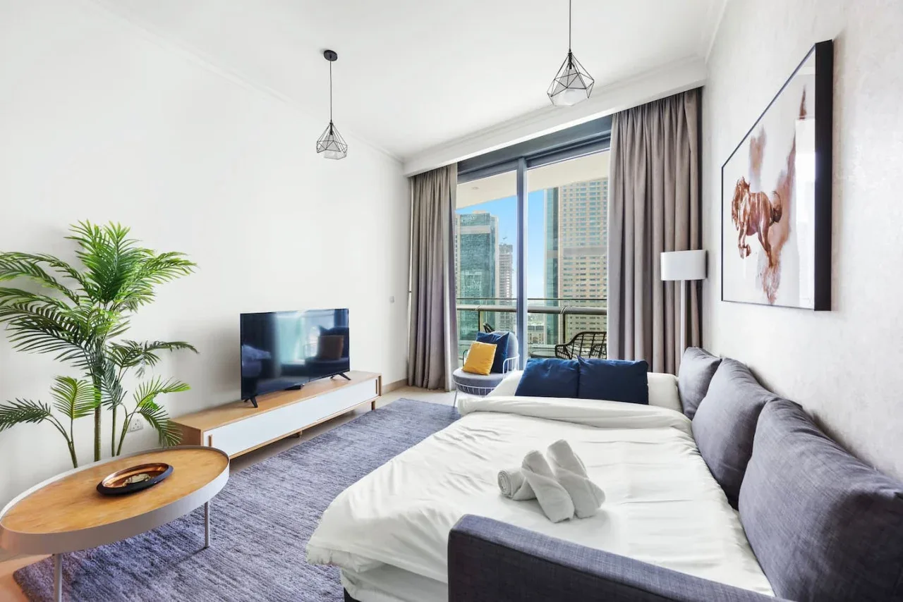 Una sala luminosa con una cama, asientos y grandes ventanas con vistas a edificios de la ciudad.