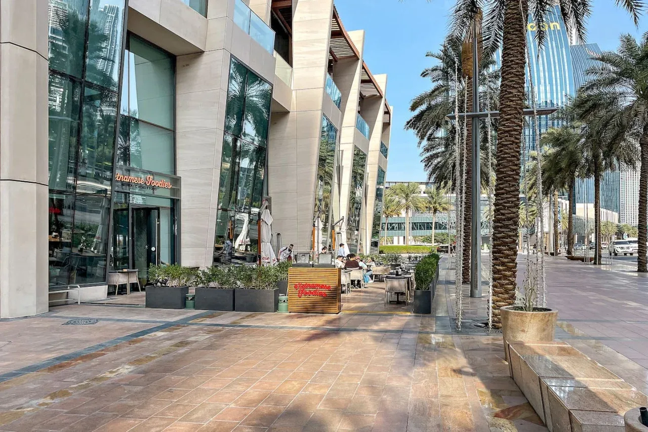Una plaza exterior pavimentada junto a un edificio moderno con fachada de vidrio, palmeras y jardineras.