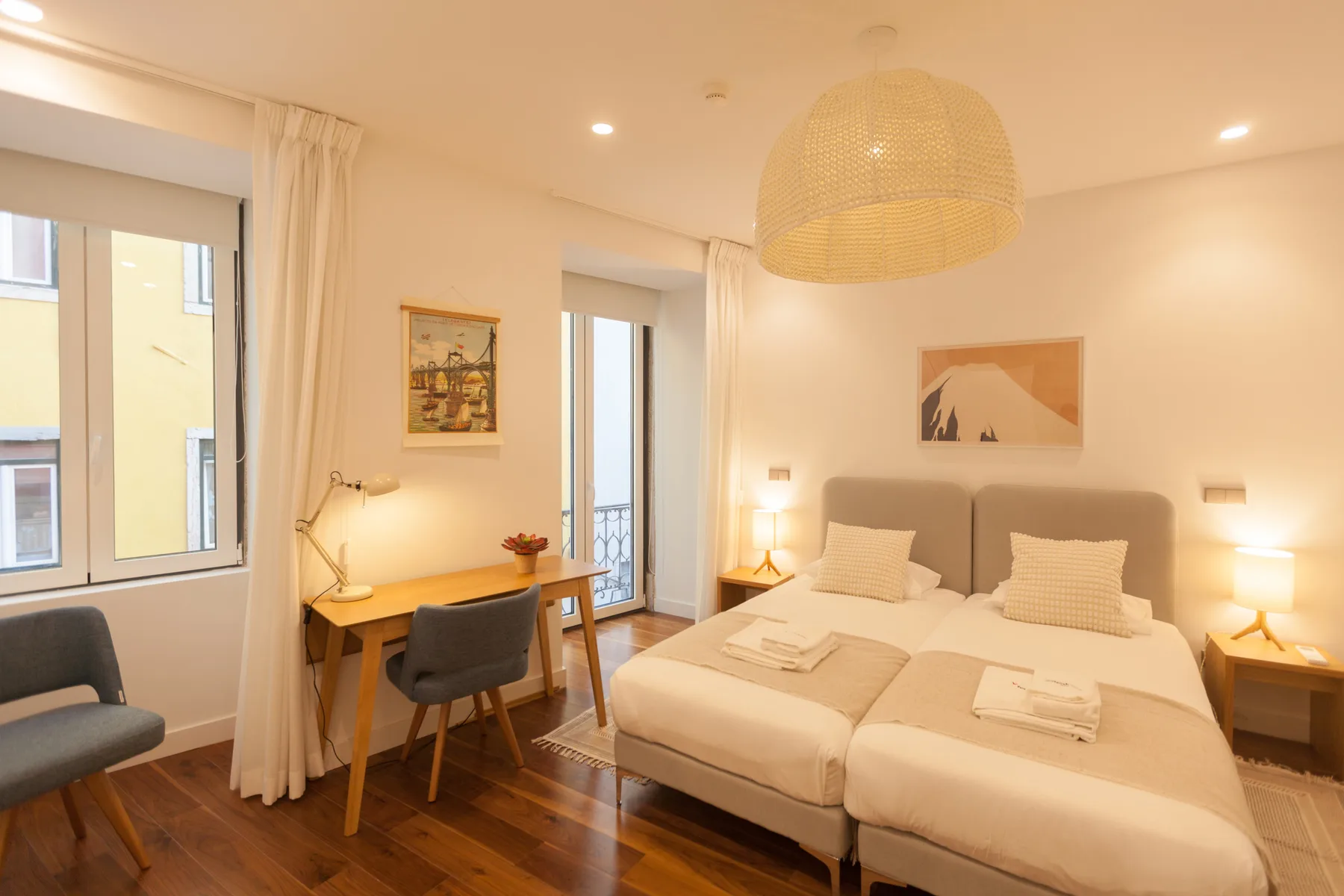 A bright bedroom with two beds, bedside tables, and a desk by the window.