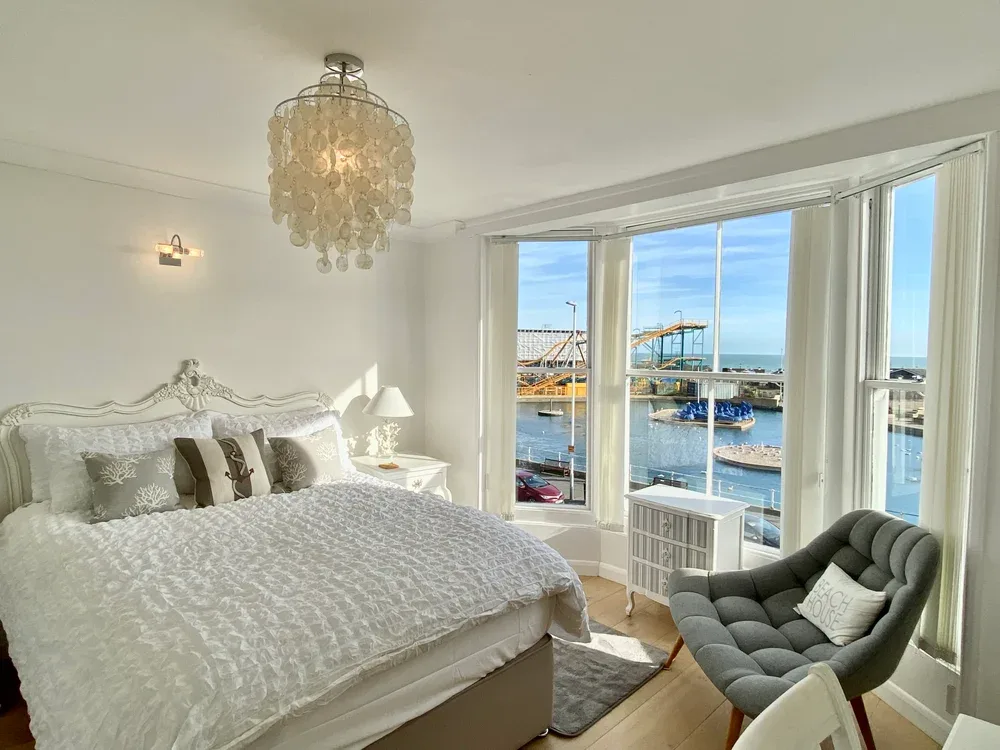 A bright bedroom with a large bed and a bay window overlooking the water.