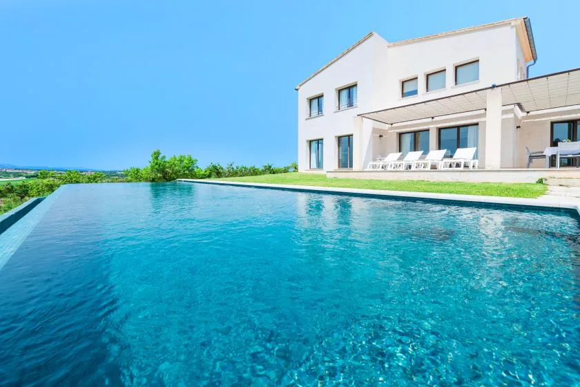 Una piscina exterior se extiende hacia una casa blanca moderna bajo un cielo despejado.