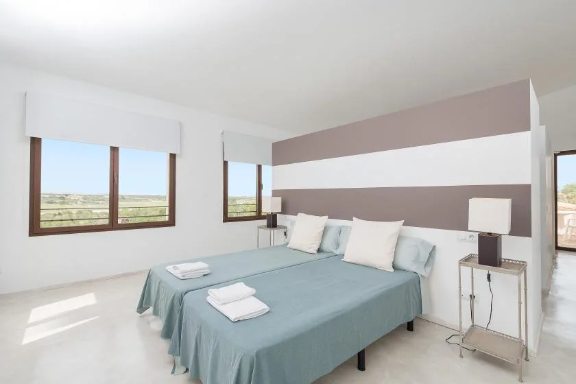 Un dormitorio luminoso con una cama vestida con ropa de cama azul claro y ventanas con vistas a un campo abierto.