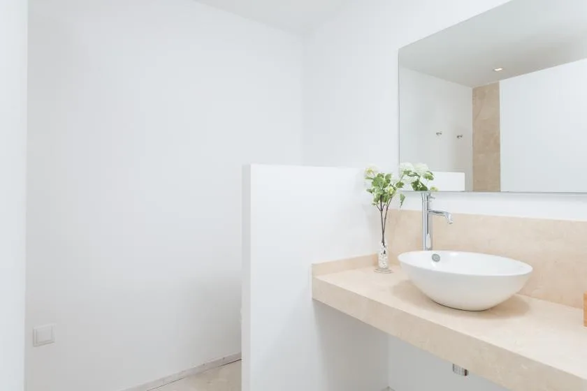 Un lavabo de baño luminoso con un lavamanos sobre encimera y un pequeño jarrón con flores.