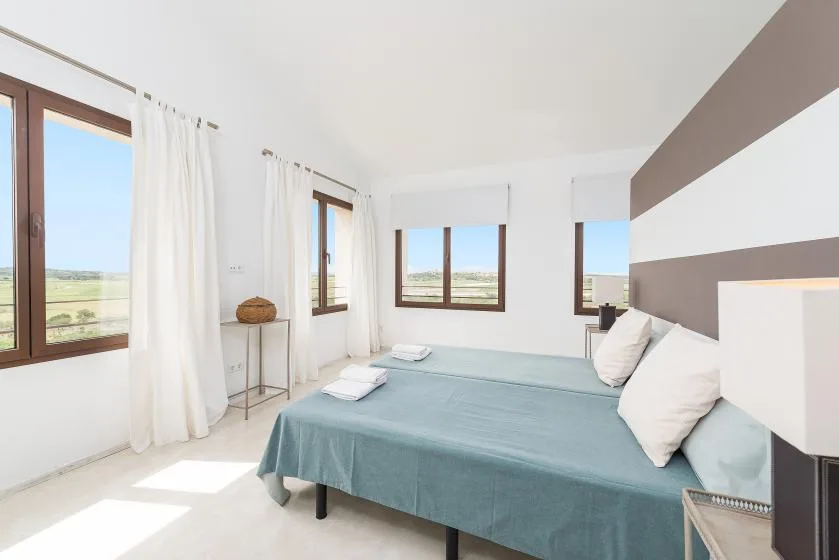 Un dormitorio luminoso con una cama, cortinas blancas y ventanas con vistas a campos abiertos.