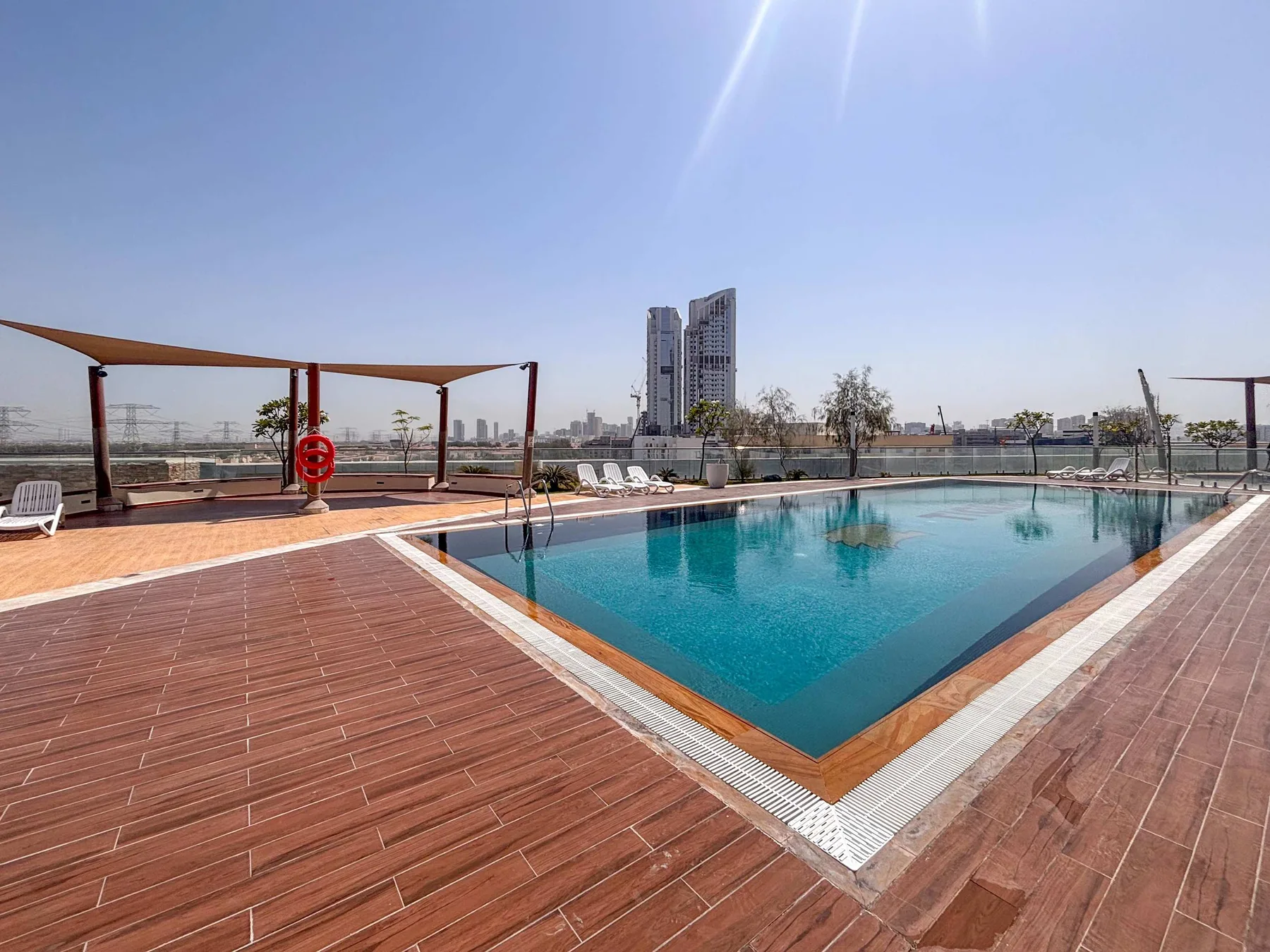 A rooftop swimming pool is surrounded by a broad wooden deck and shaded seating.