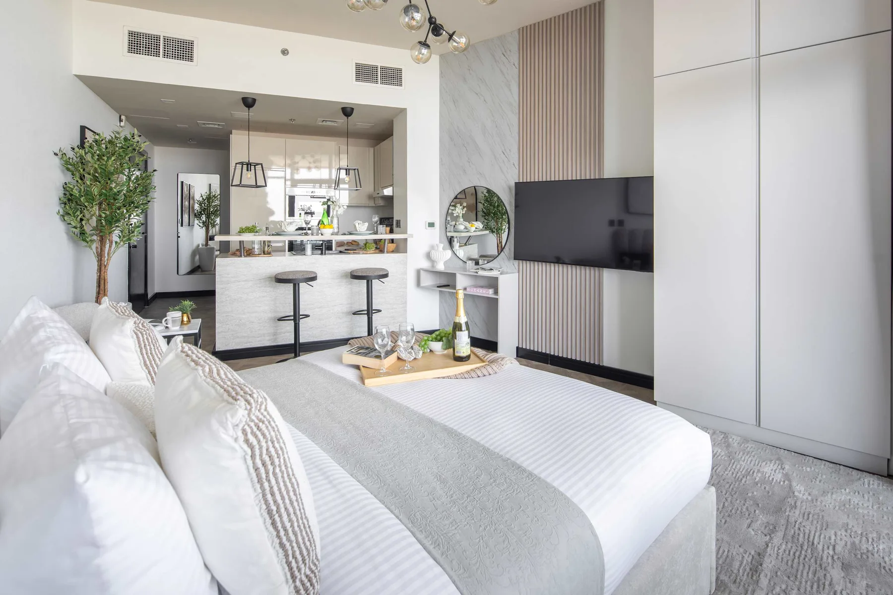 A bright bedroom with a bed facing a television and an open kitchen beyond.