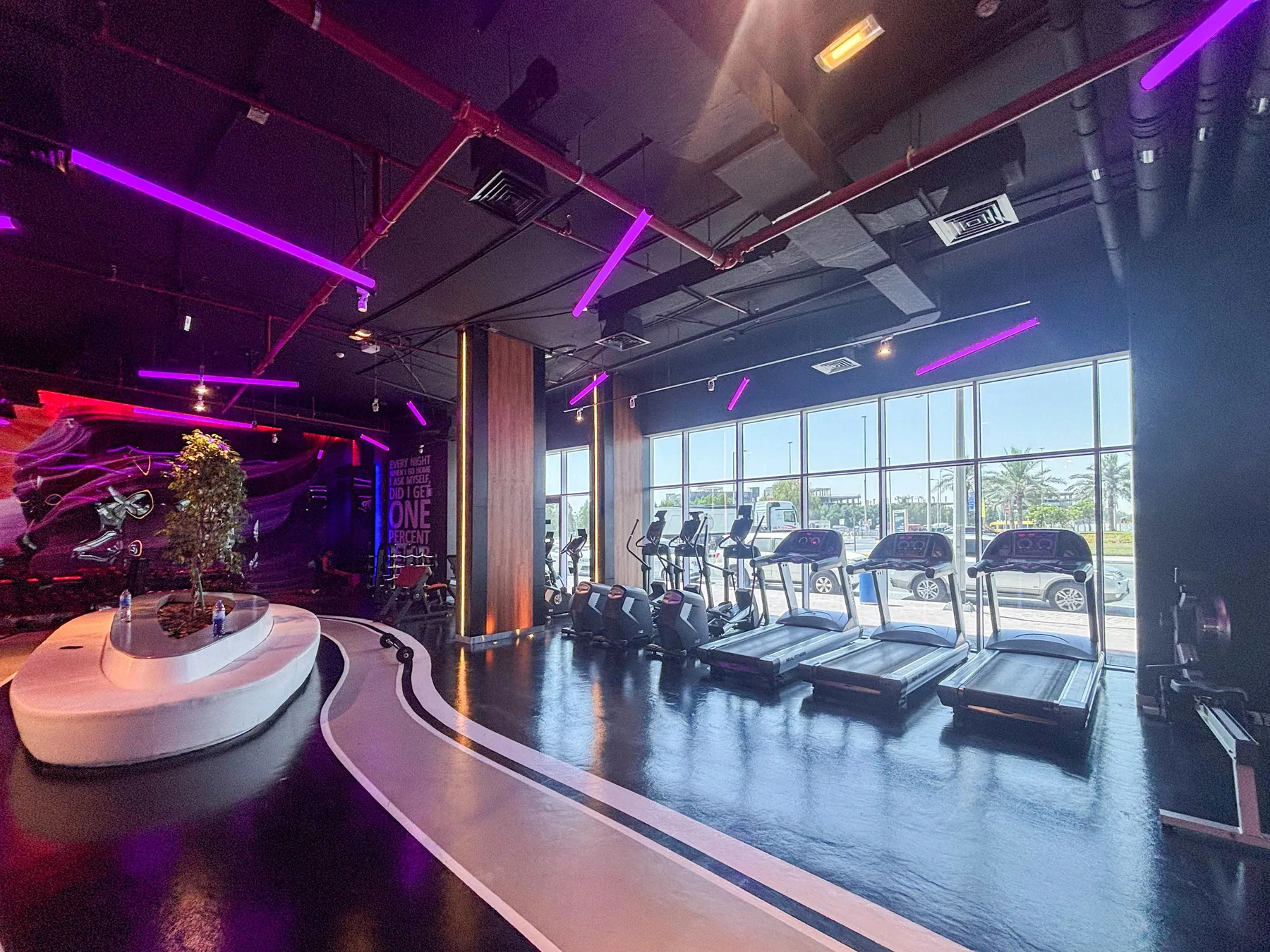 A modern gym with treadmills beside large windows and purple lighting.