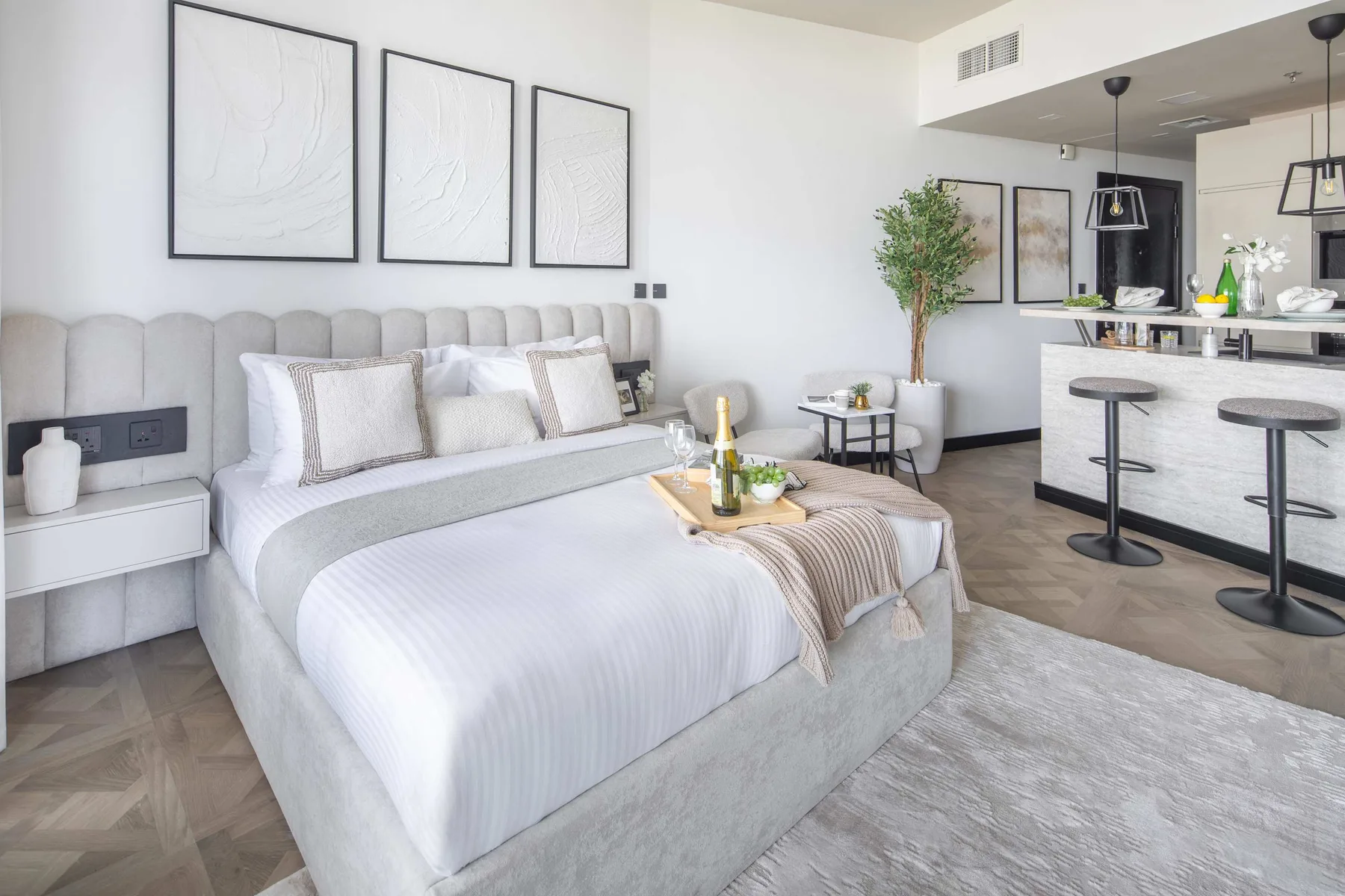 A bright bedroom with a neatly made bed and a kitchen counter in the background.