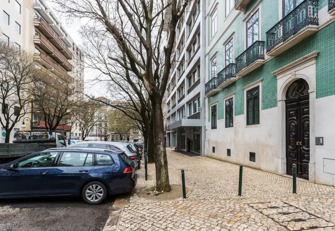 Una calle urbana con coches aparcados, árboles y un edificio con azulejos verdes.