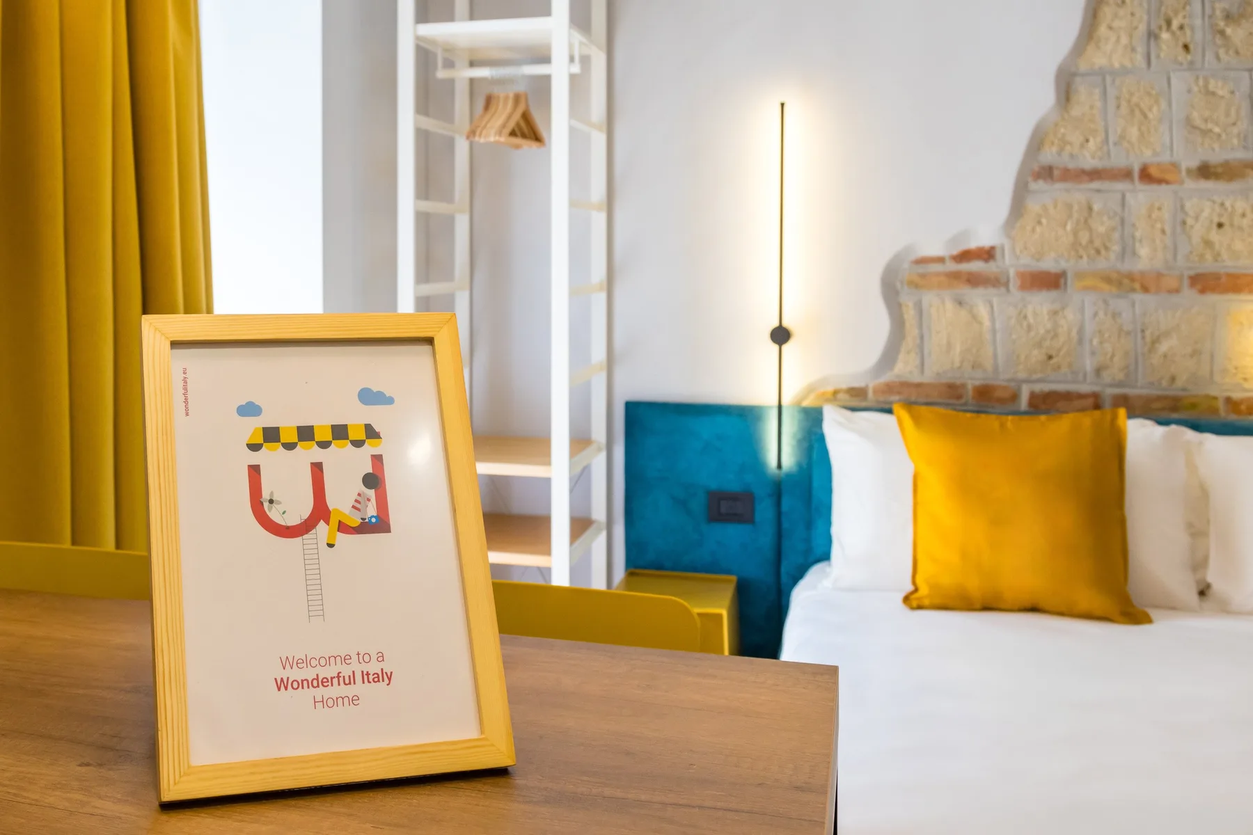 Un dormitorio con una cama, cojines de colores y una lámina de bienvenida enmarcada sobre una mesa.