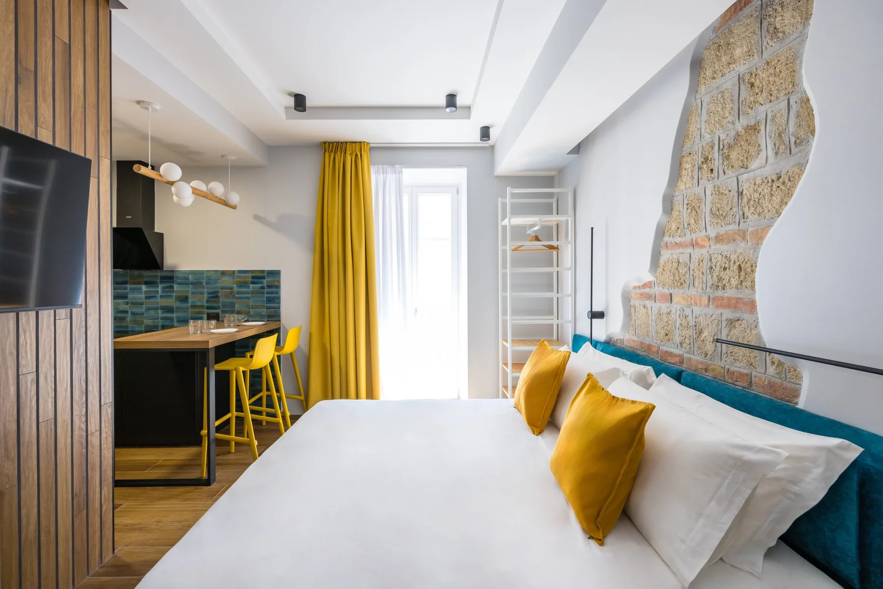 Un dormitorio con una cama junto a una pequeña cocina y taburetes amarillos.