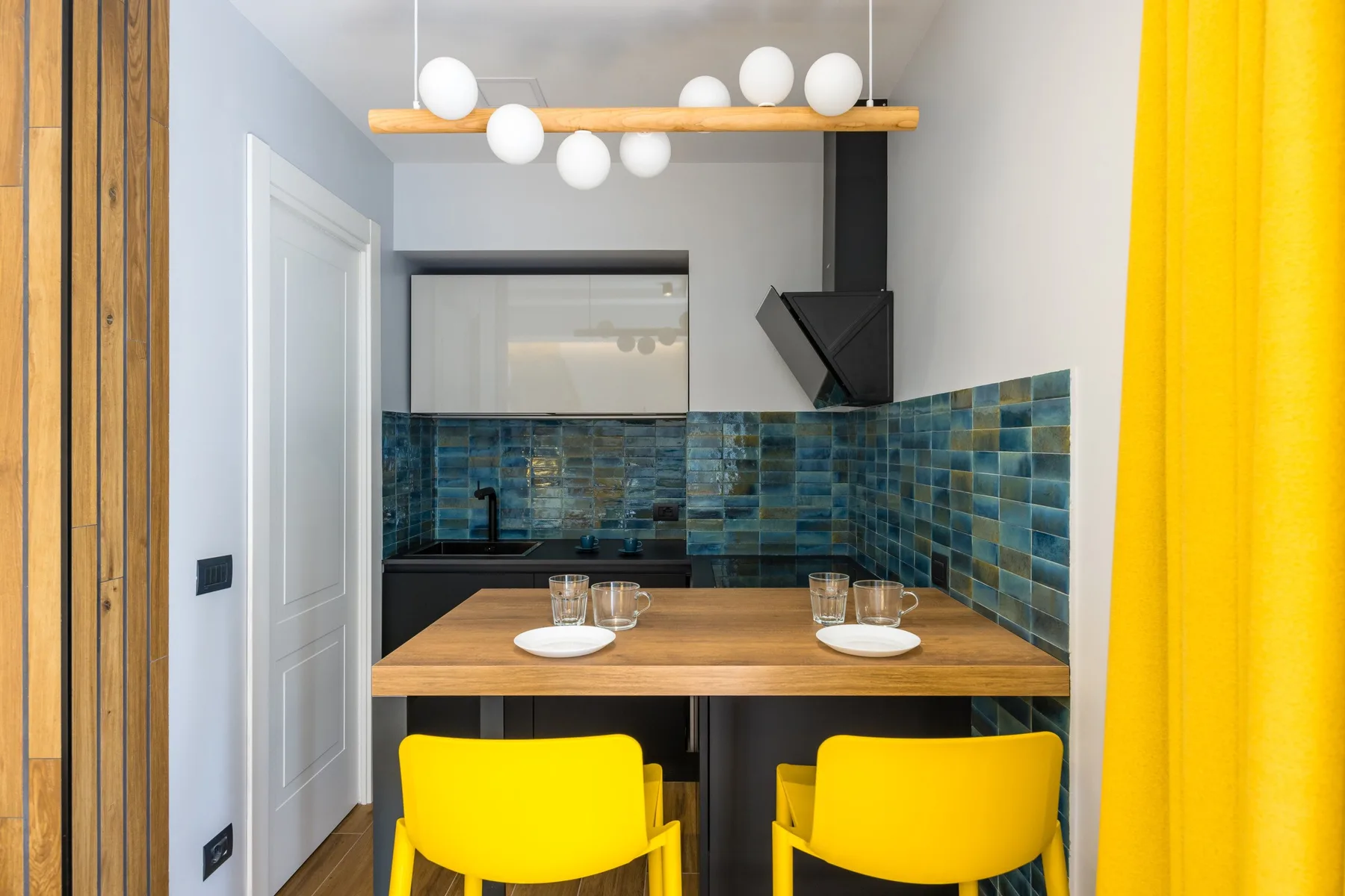 Una pequeña zona de cocina y comedor tiene una mesa de madera, asientos amarillos y paredes de azulejos azules.