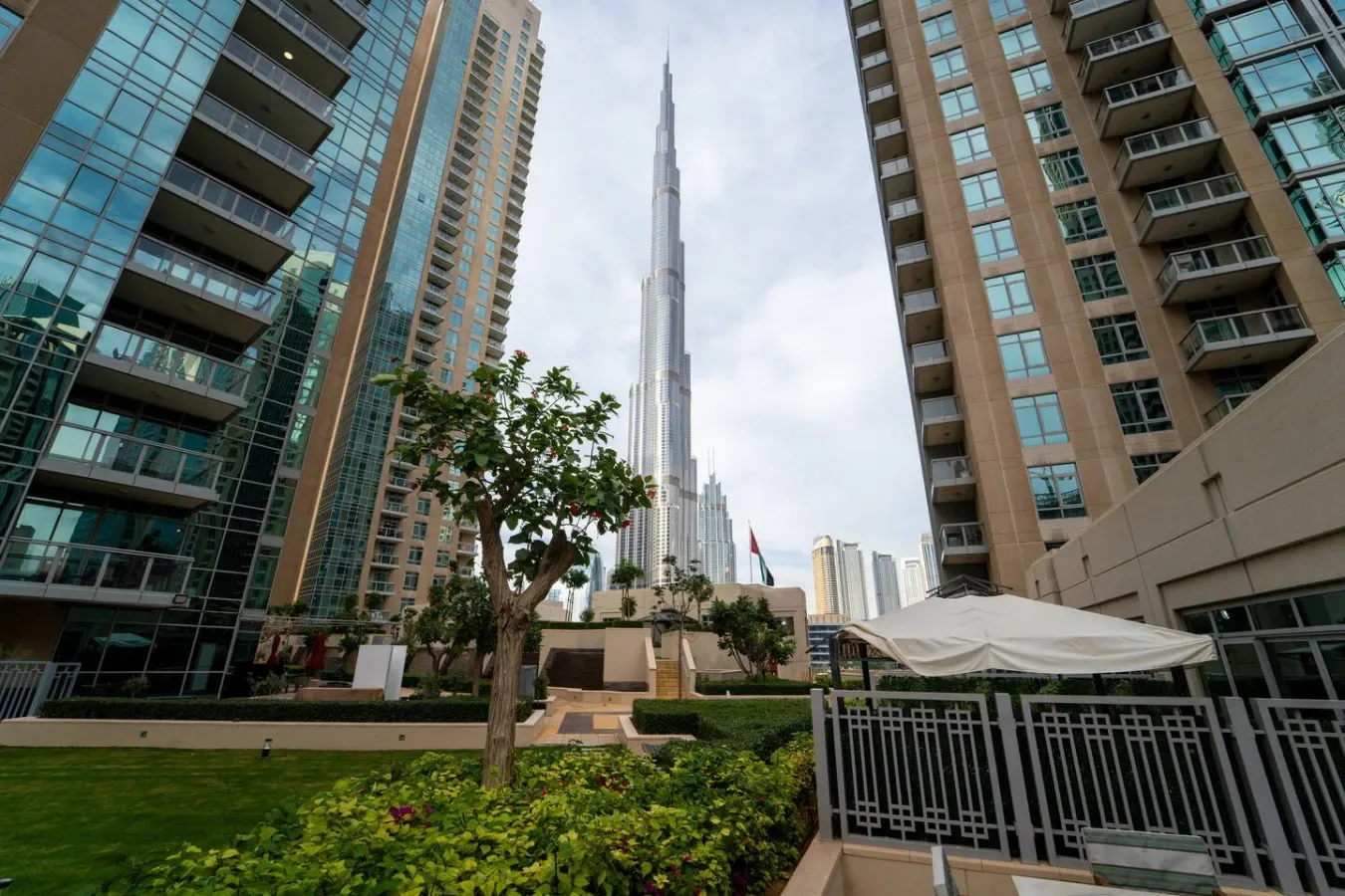 Un patio ajardinado se encuentra entre edificios altos, con una torre elevada al fondo.