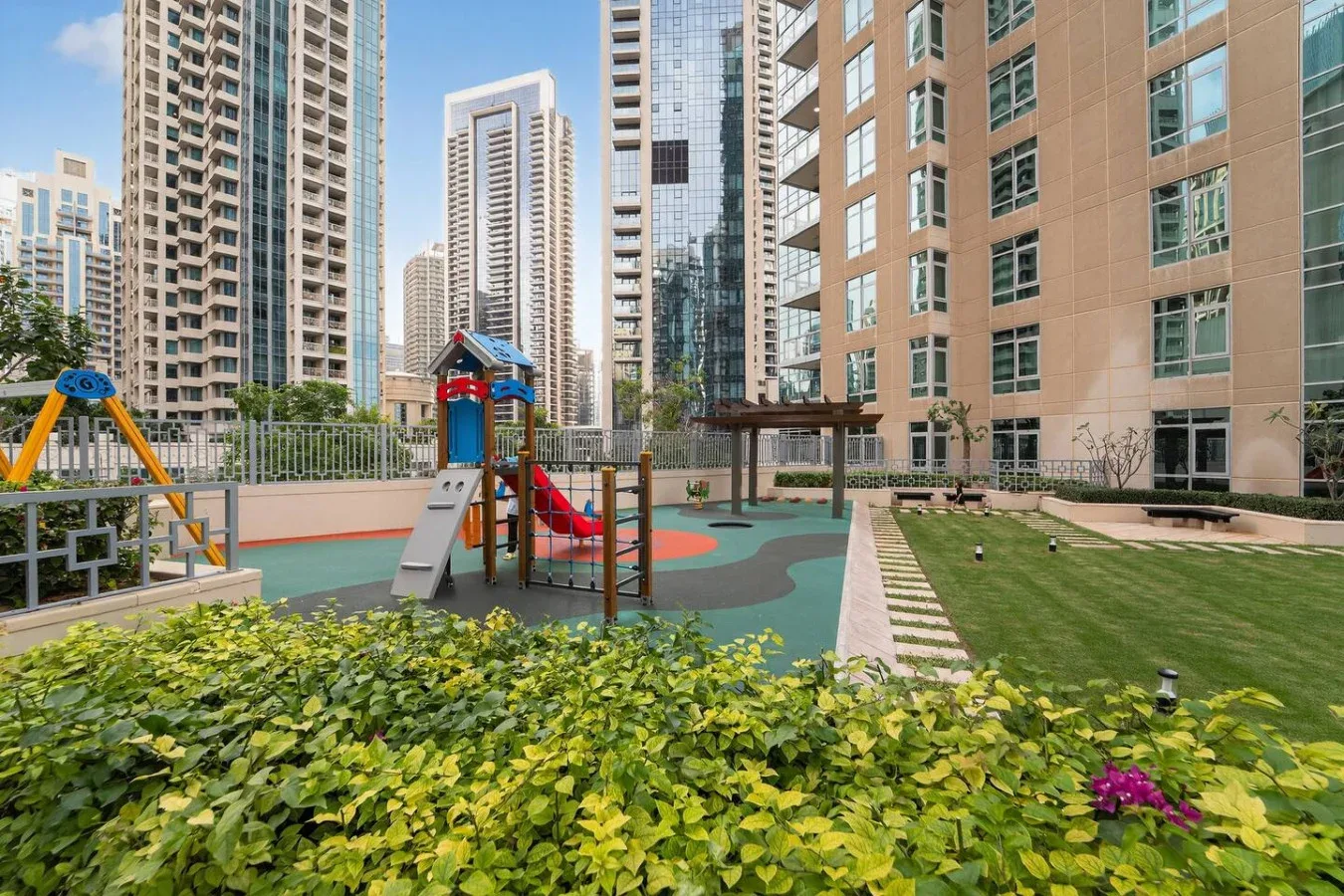 Un parque infantil ajardinado está entre edificios residenciales altos, con juegos y césped.