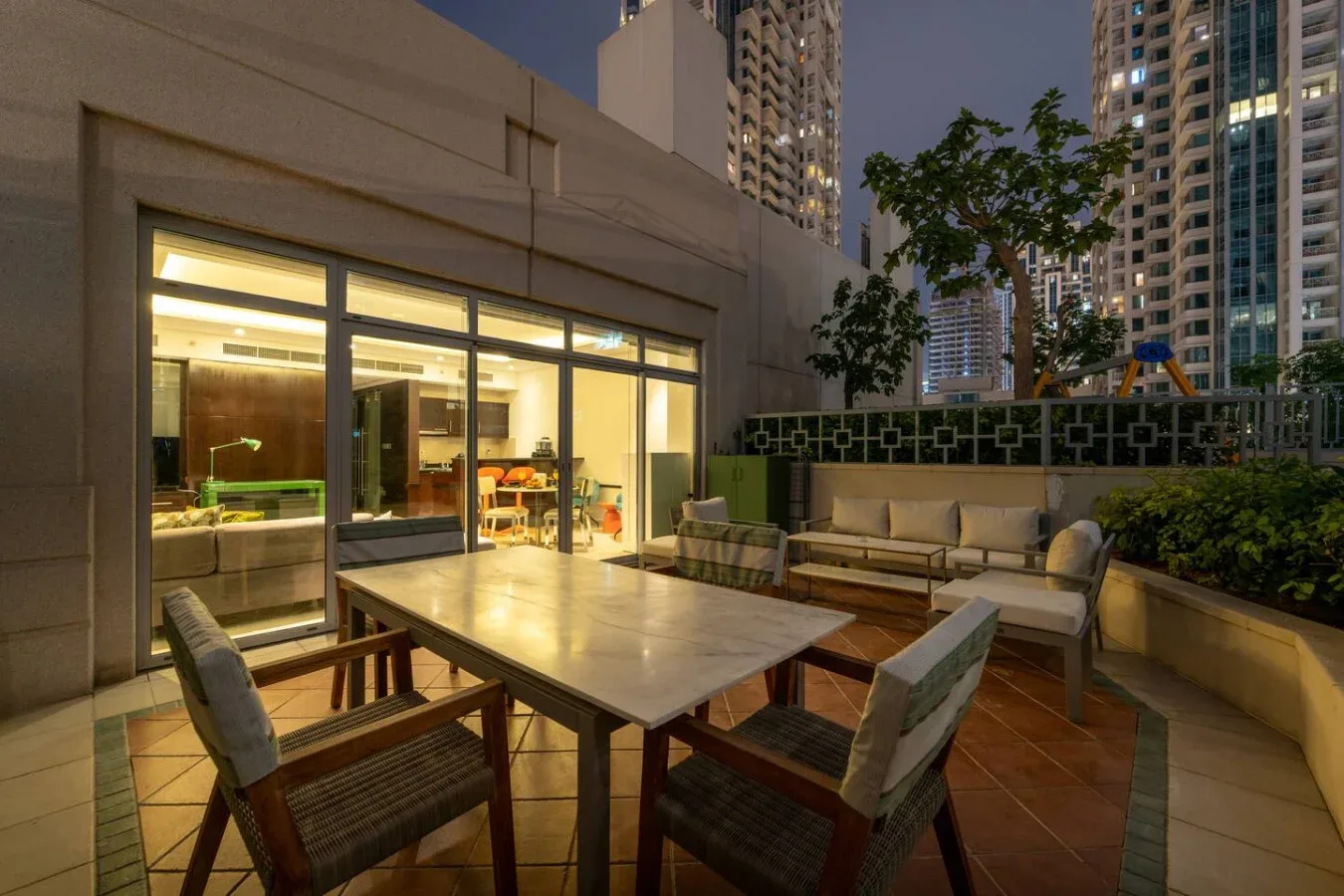 Un patio exterior con mesa y asientos junto a ventanas iluminadas.