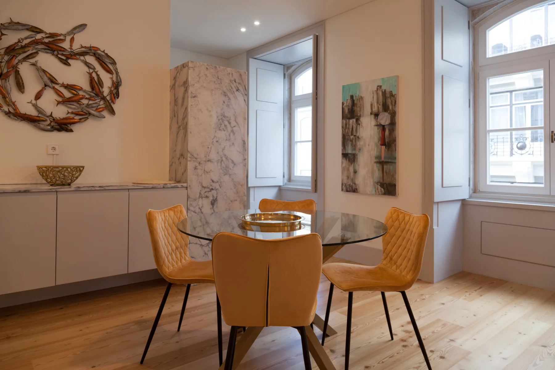 Dining area with a round table and orange seating beside tall windows.