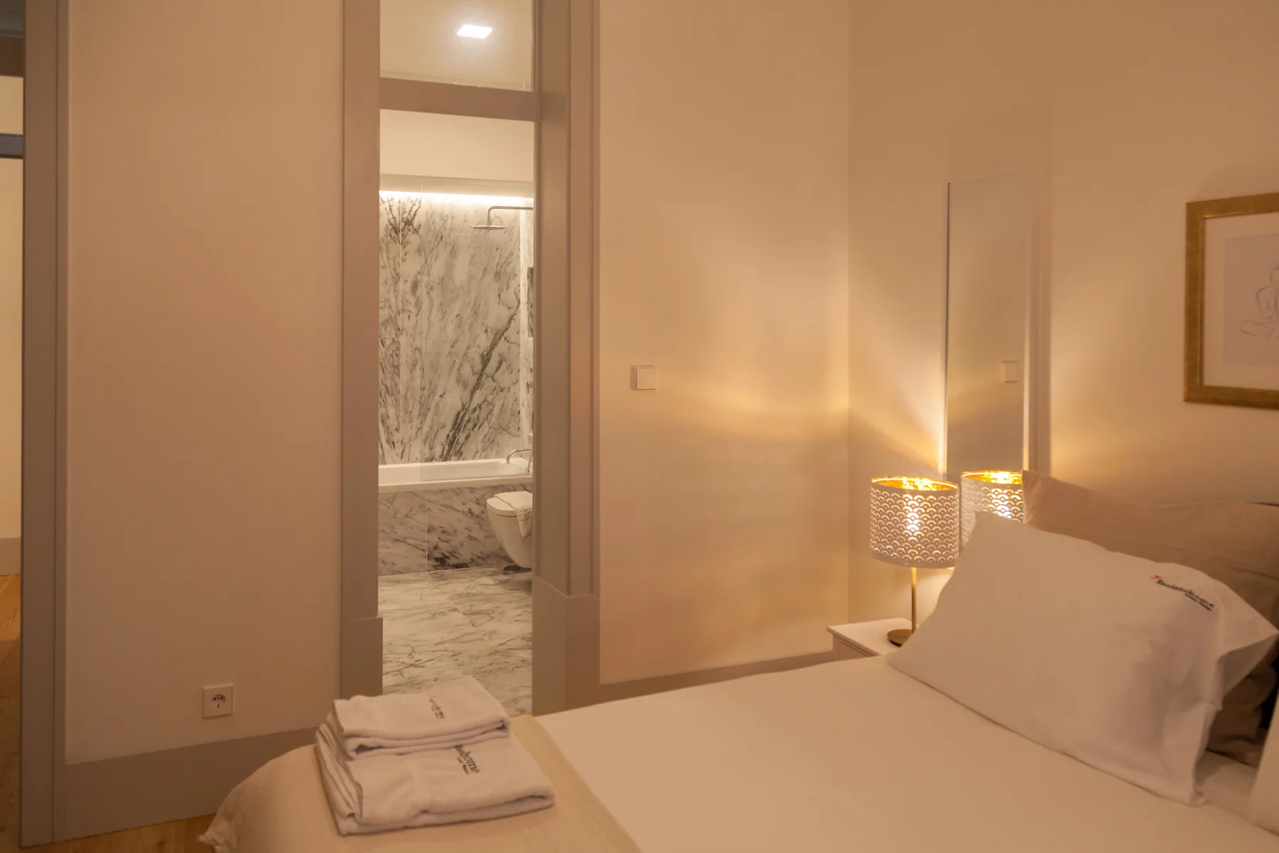 A bedroom with a neatly made bed and an open doorway to a marble bathroom.