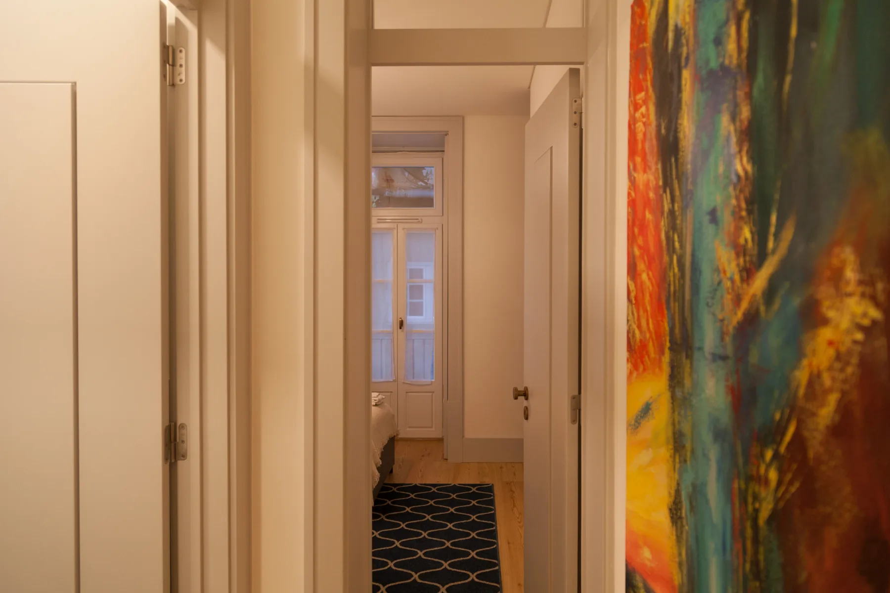 A hallway leads toward a bright window, with a colorful painting along the wall.