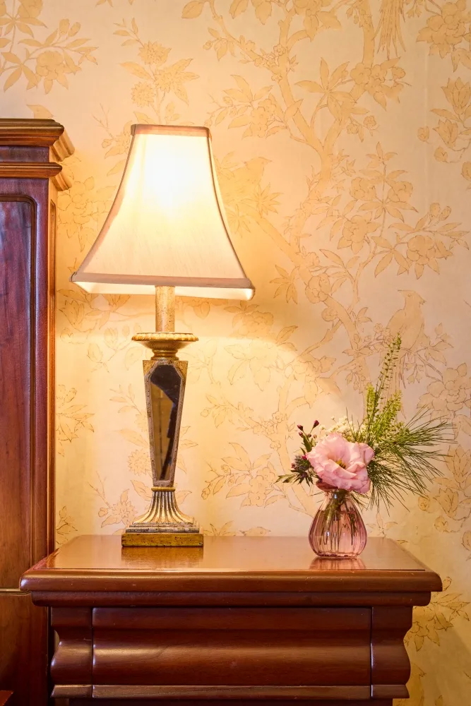 Bedside table with a lamp and flower against floral wallpaper.