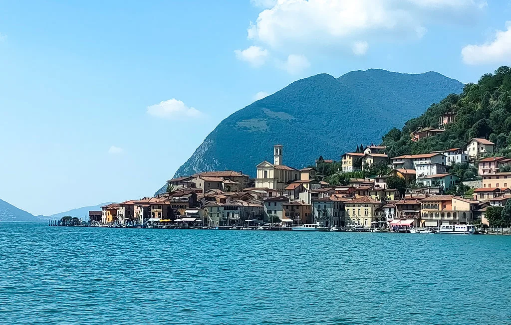 A lakeside village sits below a green mountain beside blue water.