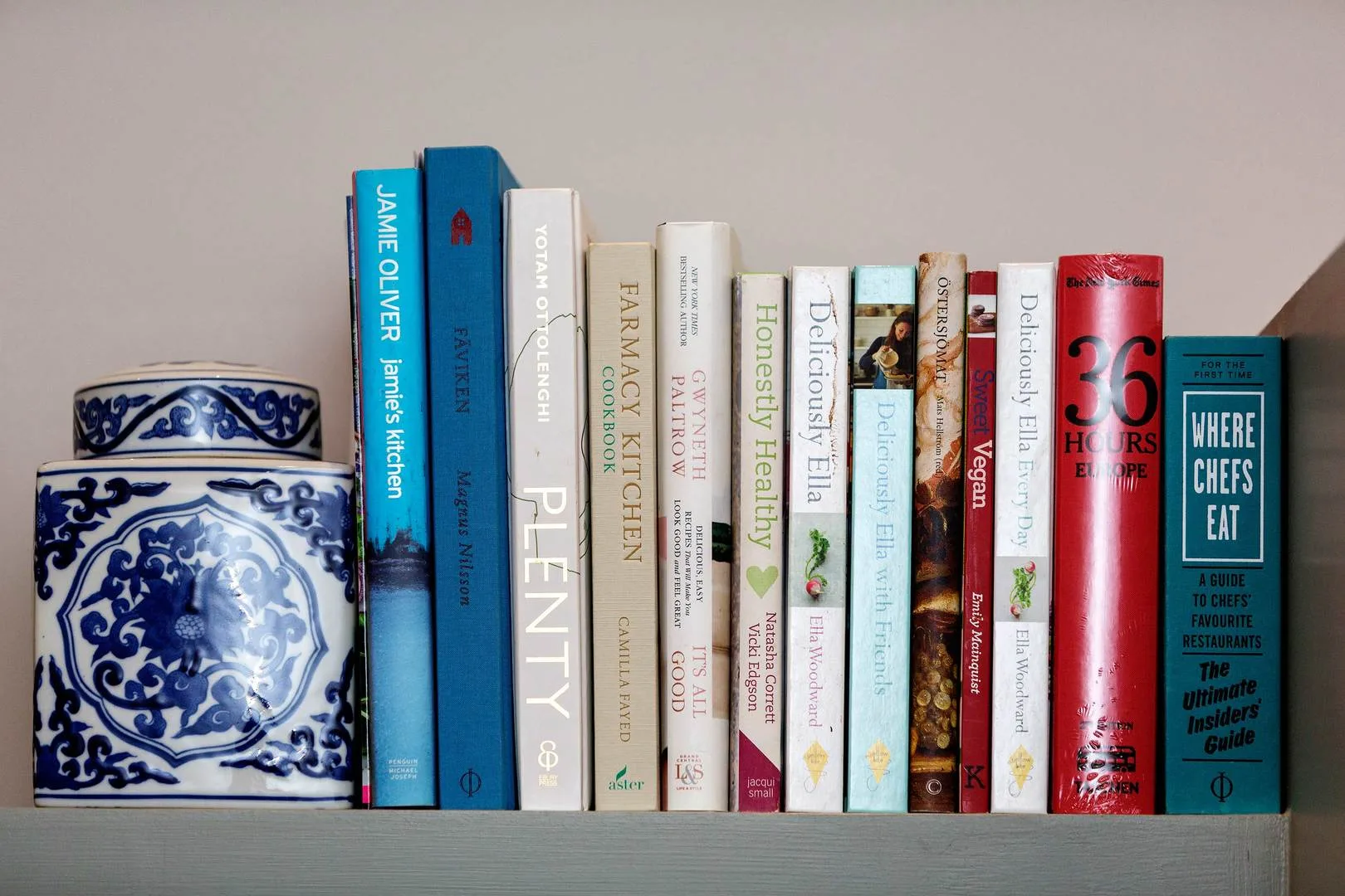 Una fila de libros de cocina está junto a un frasco decorativo azul y blanco en un estante.