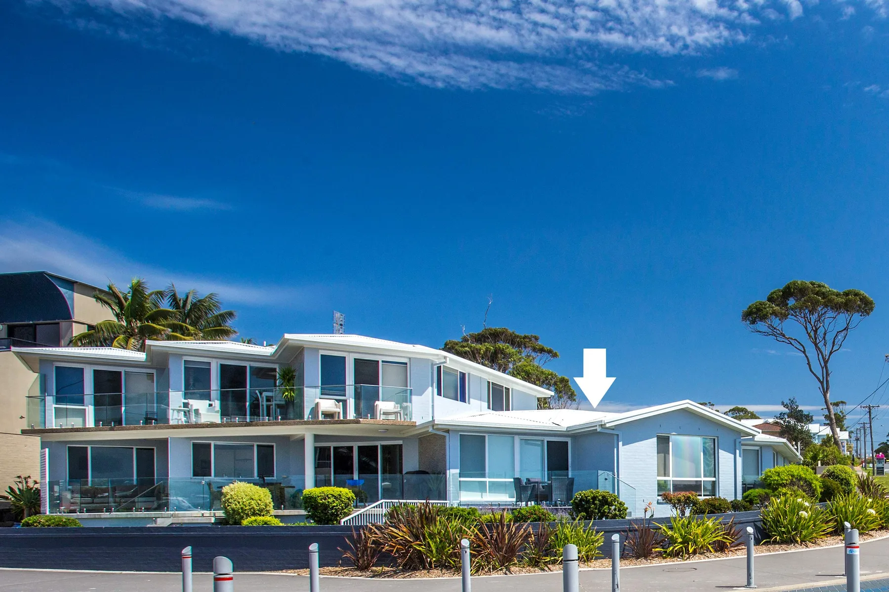Edificios modernos junto al mar, con balcones acristalados y arbustos bajo un cielo azul.