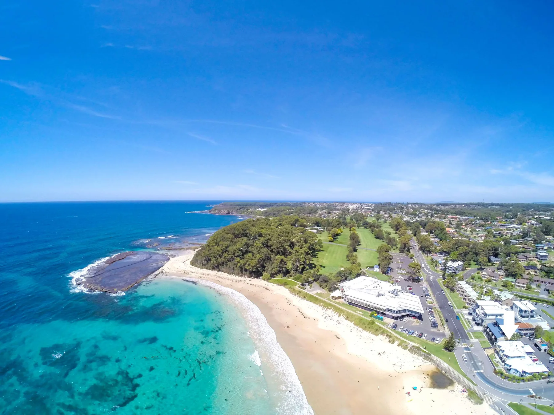 Vista aérea de una playa curva, aguas turquesas y edificios costeros cercanos.