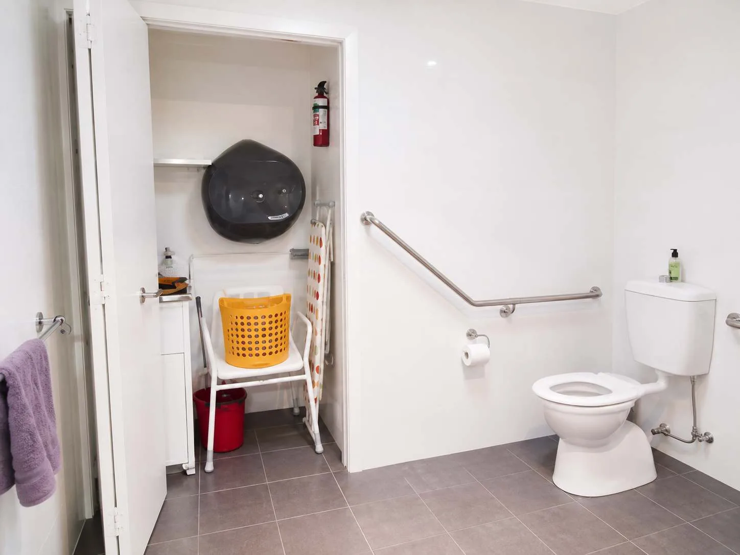 Un baño con inodoro, barra de apoyo en la pared y un espacio de almacenamiento abierto.