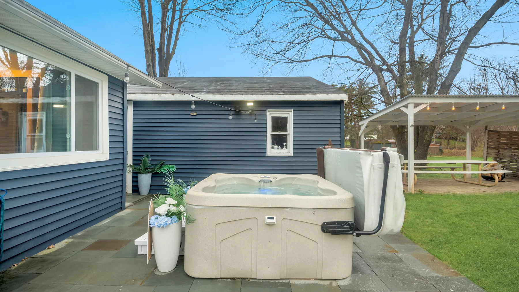 Una bañera de hidromasaje exterior está junto a una casa azul y un patio cubierto.