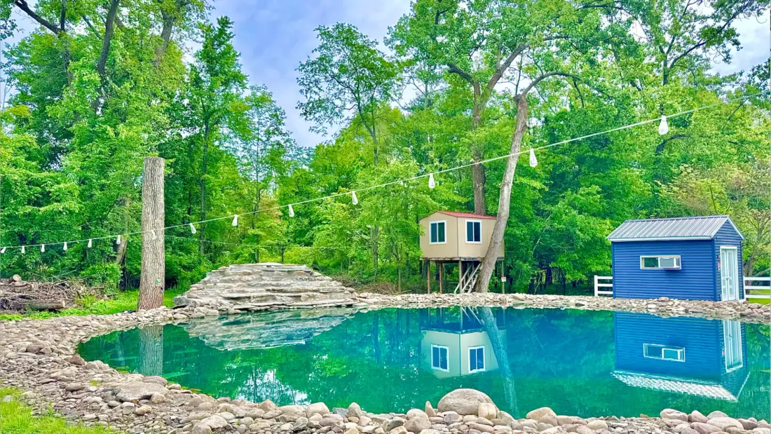Un estanque de baño al aire libre está junto a una pequeña estructura elevada y un cobertizo.