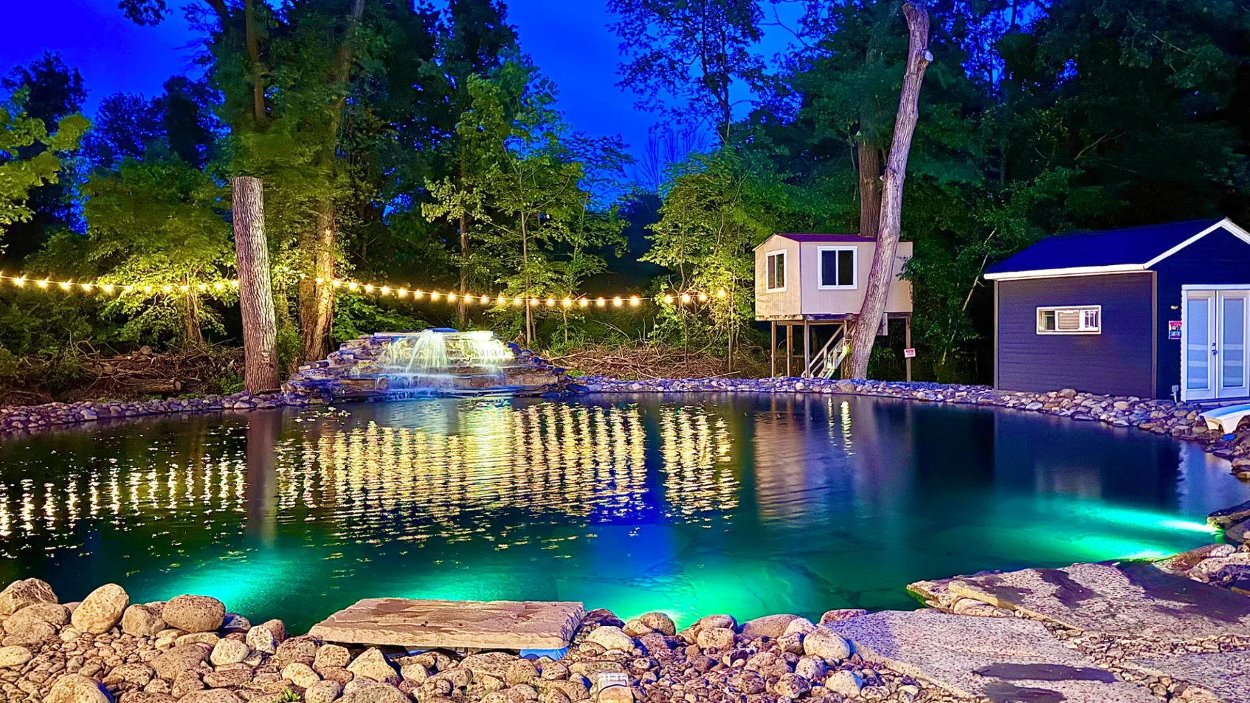 Una piscina exterior iluminada está entre árboles, junto a una pequeña construcción al anochecer.