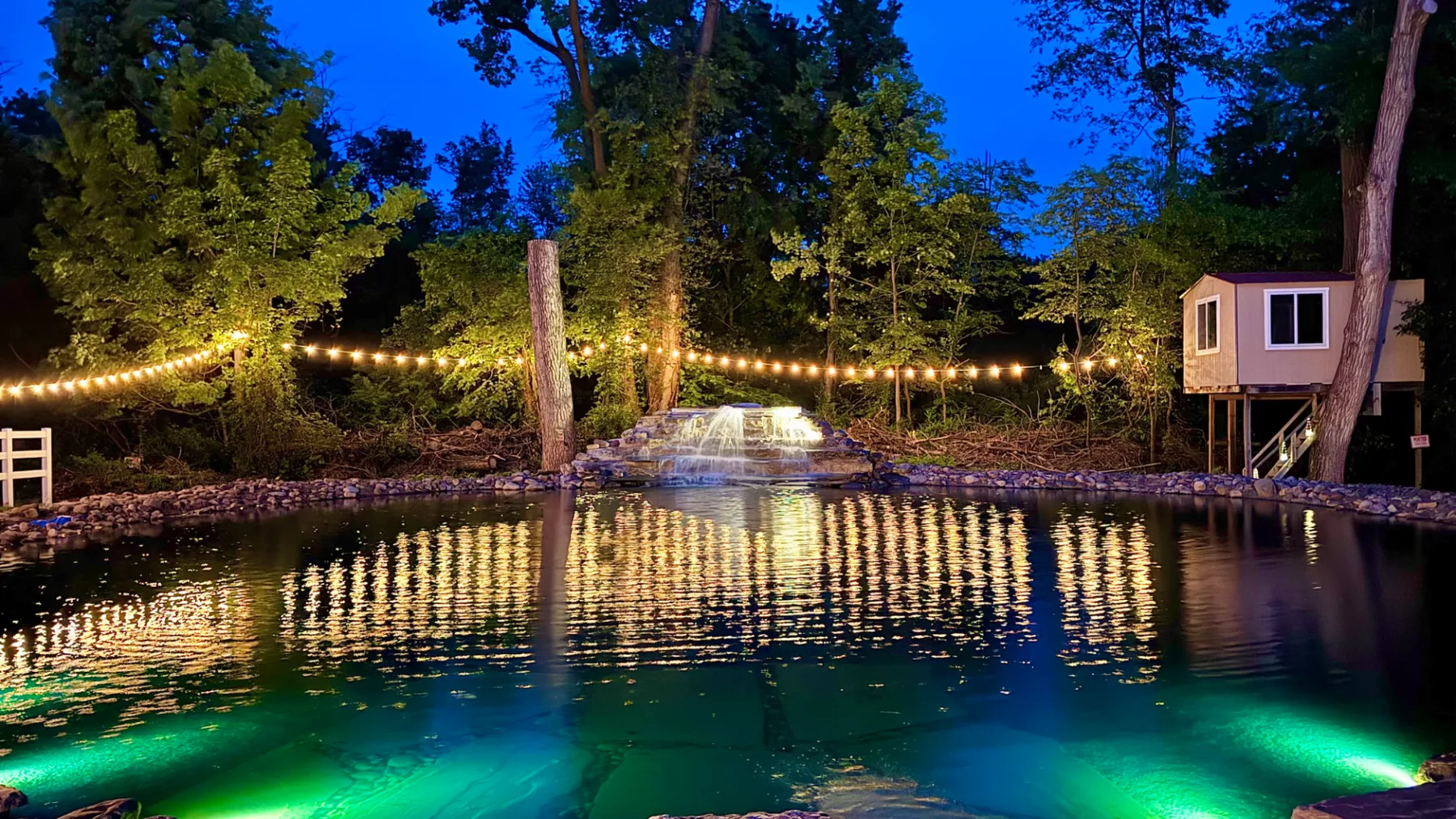 Un estanque iluminado, rodeado de árboles y luces colgantes al anochecer.