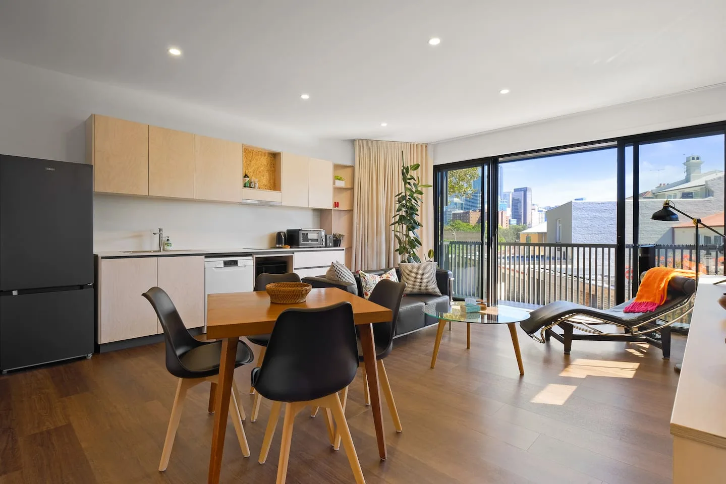 An open-plan dining and kitchen area has a wooden table beside large glass doors opening onto a balcony.