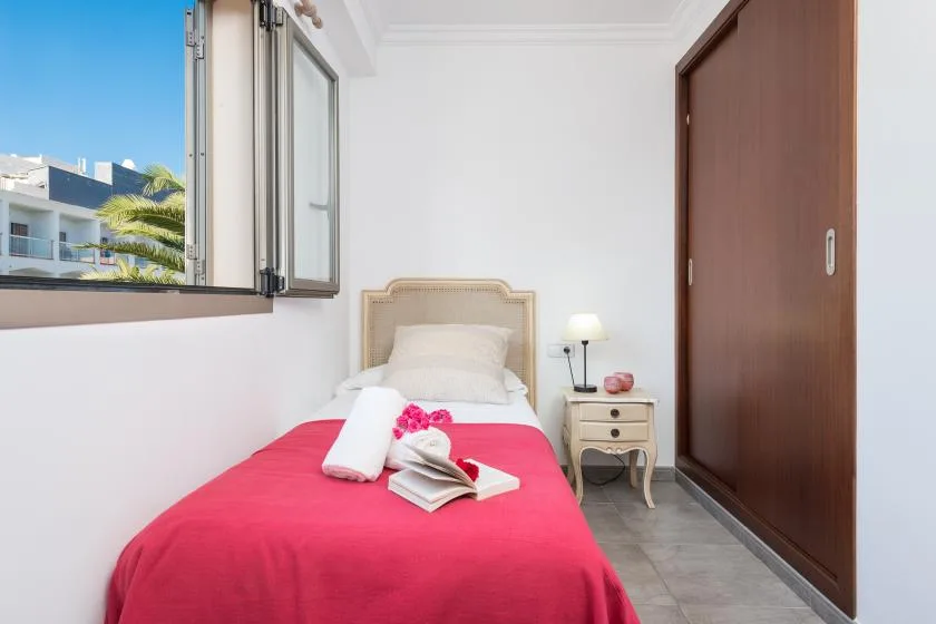 A bedroom with a bed covered by a bright red bedspread beside an open window.