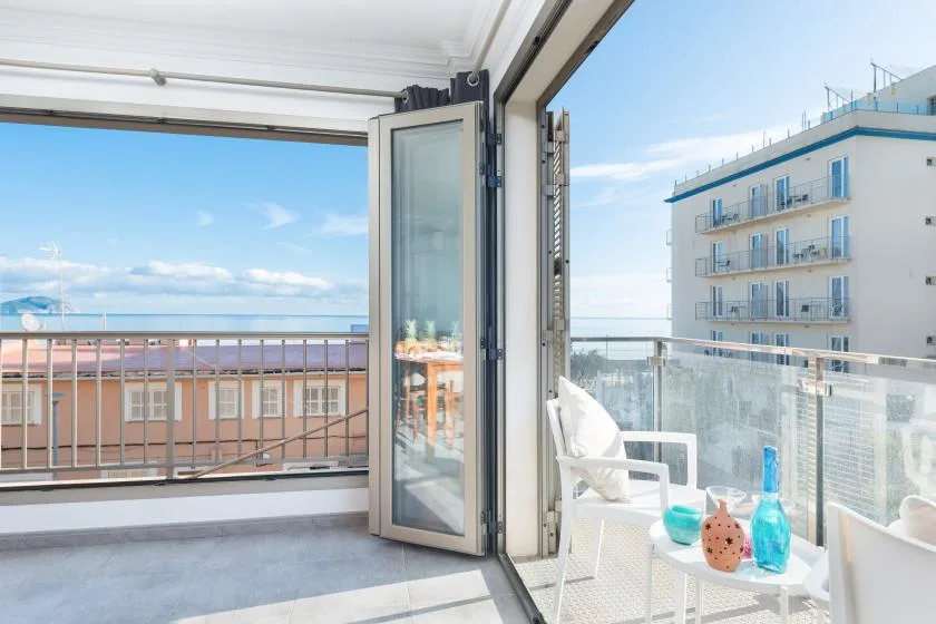 A bright balcony with glass doors, seating, and a view of nearby buildings and the sea.
