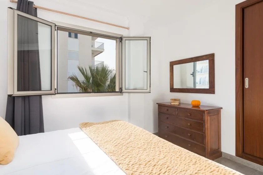 A bedroom with a bed beside an open window and a wooden dresser.
