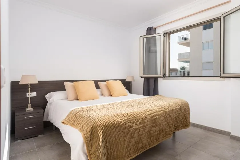 A bedroom with a bed dressed in white linens and a tan coverlet beside a window.
