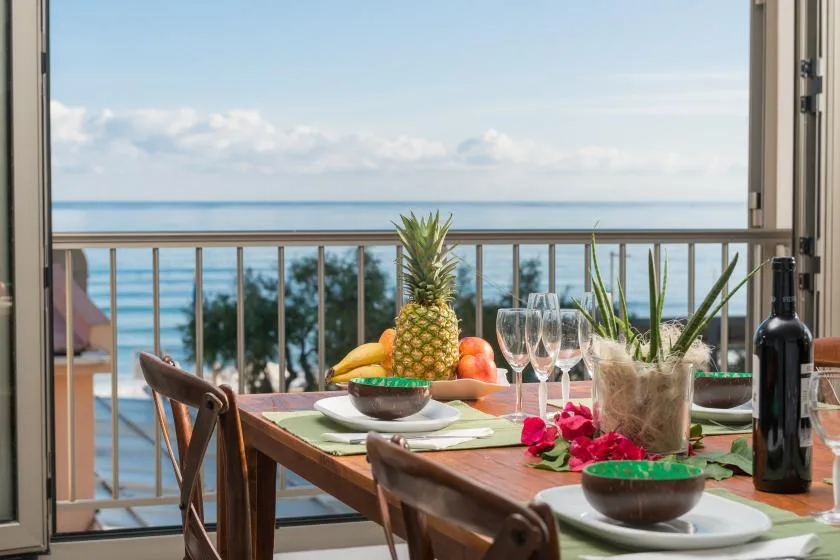 A balcony dining table with fruit and drinks overlooks the ocean.