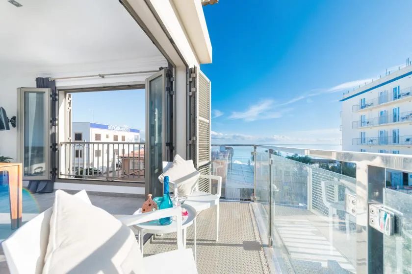 A bright balcony with seating overlooks the sea and nearby buildings.