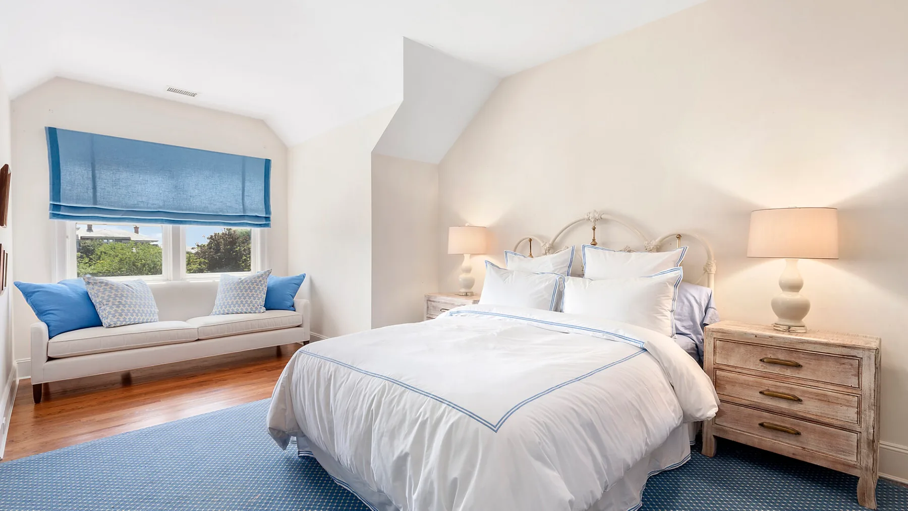 Un dormitorio luminoso con una cama bien tendida y una zona de estar junto a la ventana.