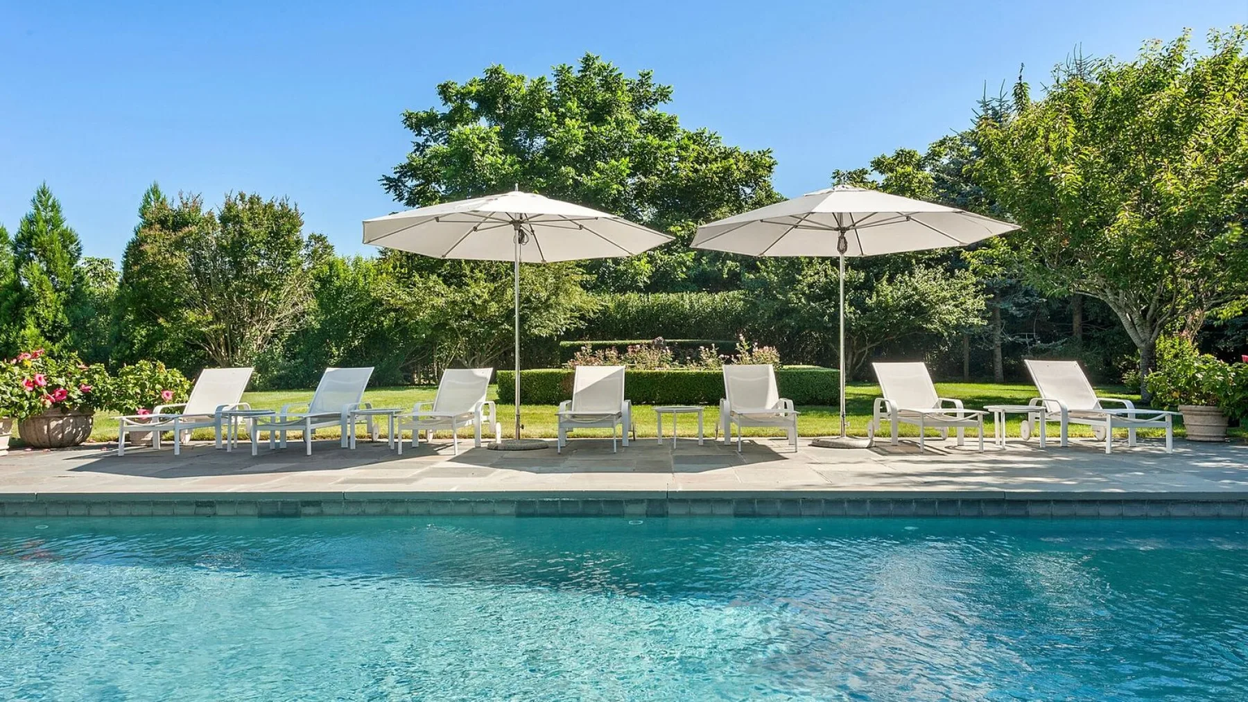 Una piscina al aire libre con asientos reclinables y sombrillas junto a un jardín frondoso.