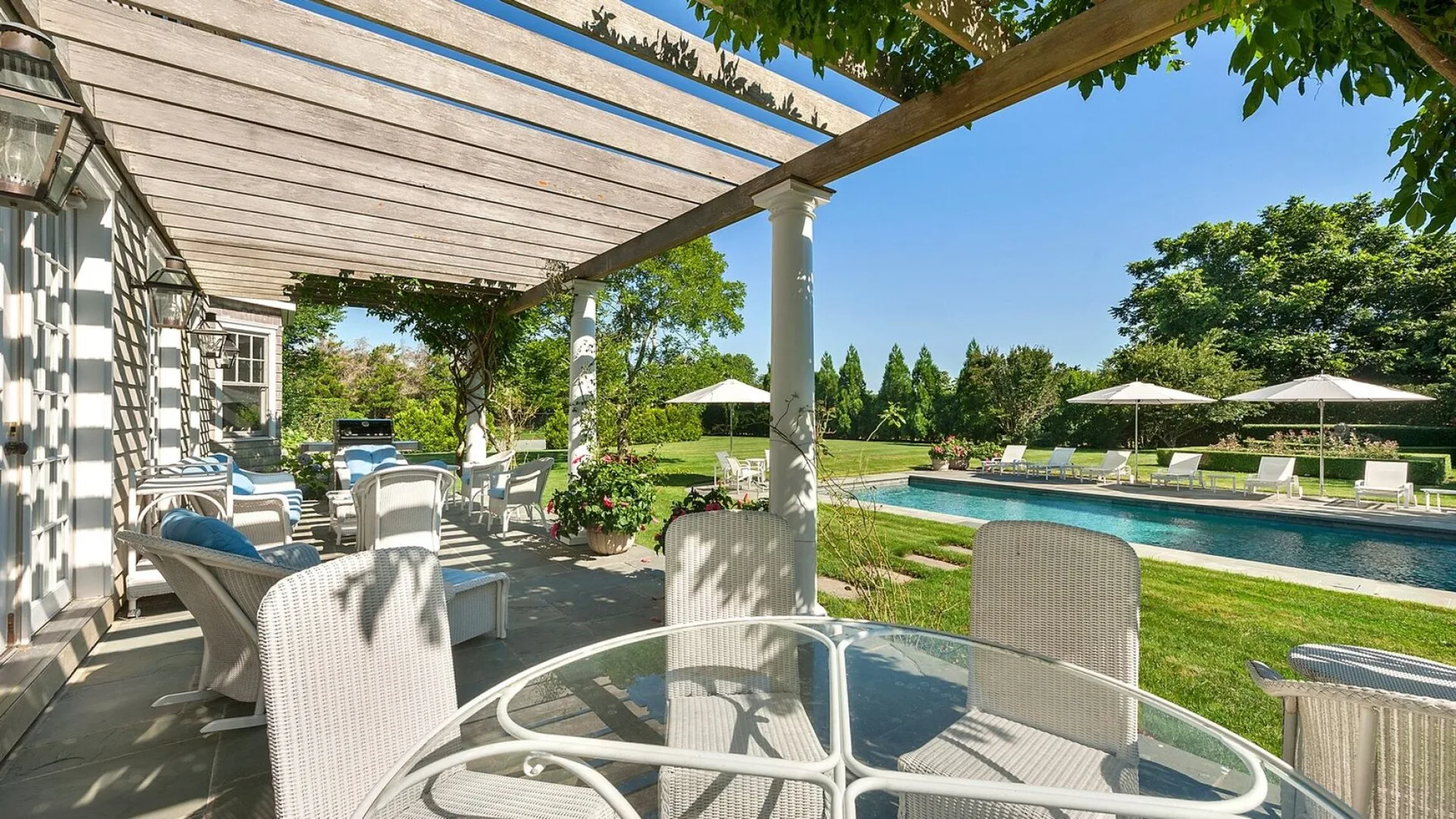 Una zona de comedor en el patio, bajo una pérgola, da a una piscina y un jardín.