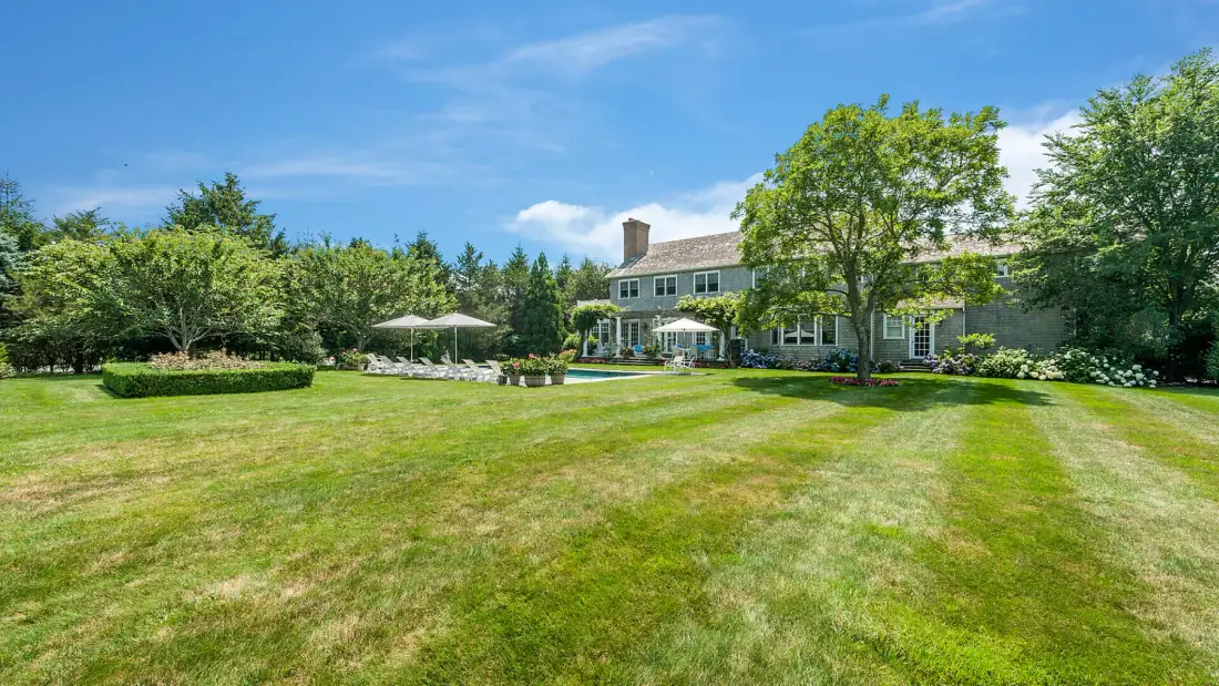 A large house stands beyond a broad green lawn, with trees and a patio umbrella.