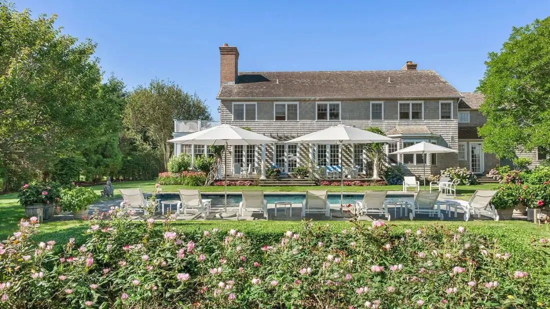 Una casa grande da a una piscina, una zona de estar al aire libre y un jardín de flores.