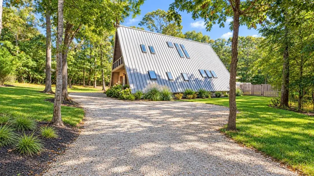 A steep-roofed house sits among trees beside a gravel driveway.