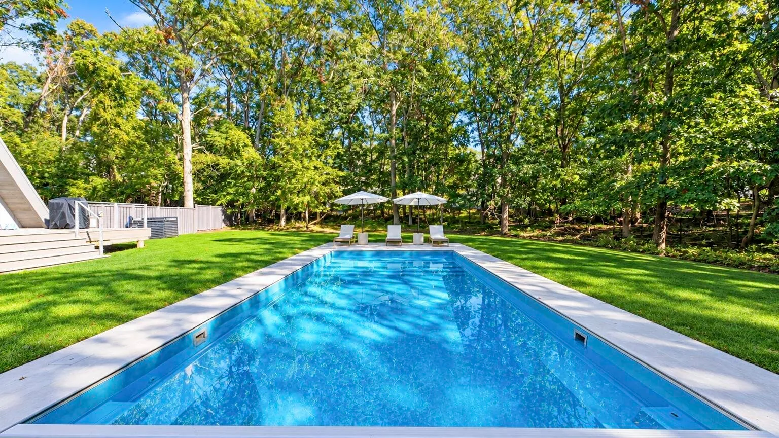 A backyard swimming pool is surrounded by green lawn and leafy trees.