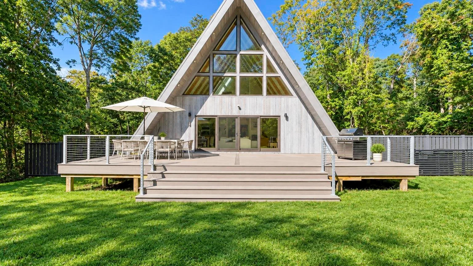 A-frame house with a broad wooden deck and outdoor seating, surrounded by trees.