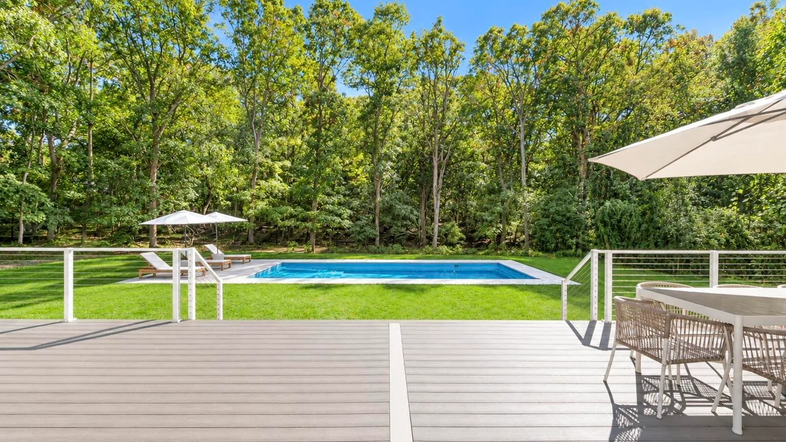 A wooden deck overlooks a swimming pool, with trees beyond the lawn.