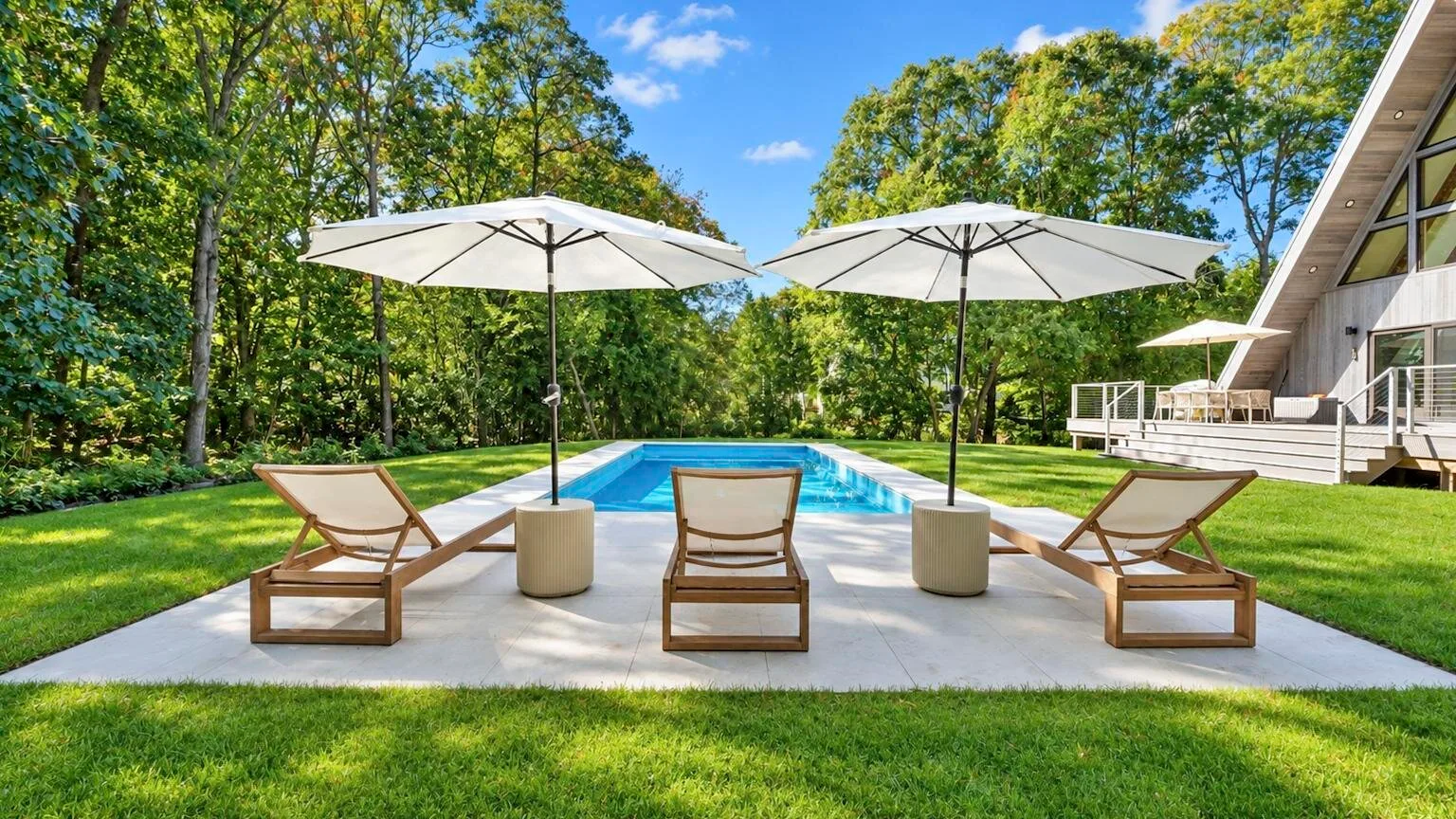 An outdoor swimming pool with lounge seating and umbrellas beside a house.