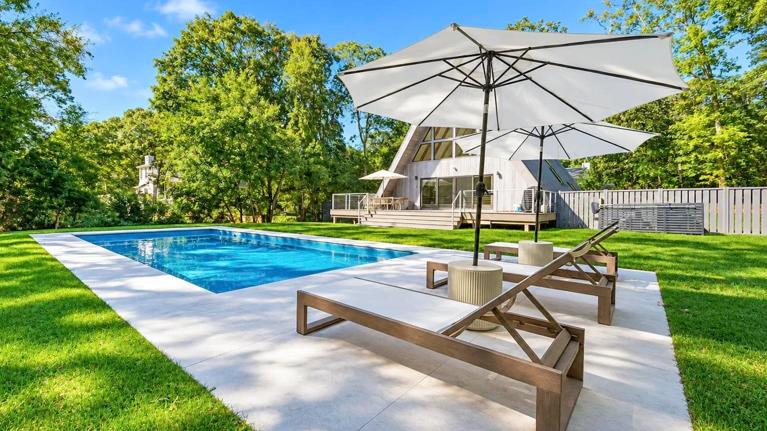 An outdoor swimming pool beside a patio with lounge seating and a large umbrella.